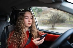 A young woman looking at a mobile phone while driving a car. A female driver in the driver's seat uses a cell phone in the auto. Girl using navigation on her smartphone. Car travel, technology. 