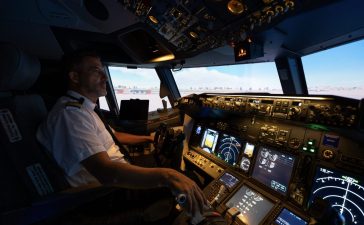 Airline pilot at the controls of a flight simulator, with illuminated cockpit instruments and an airport scene ahead.