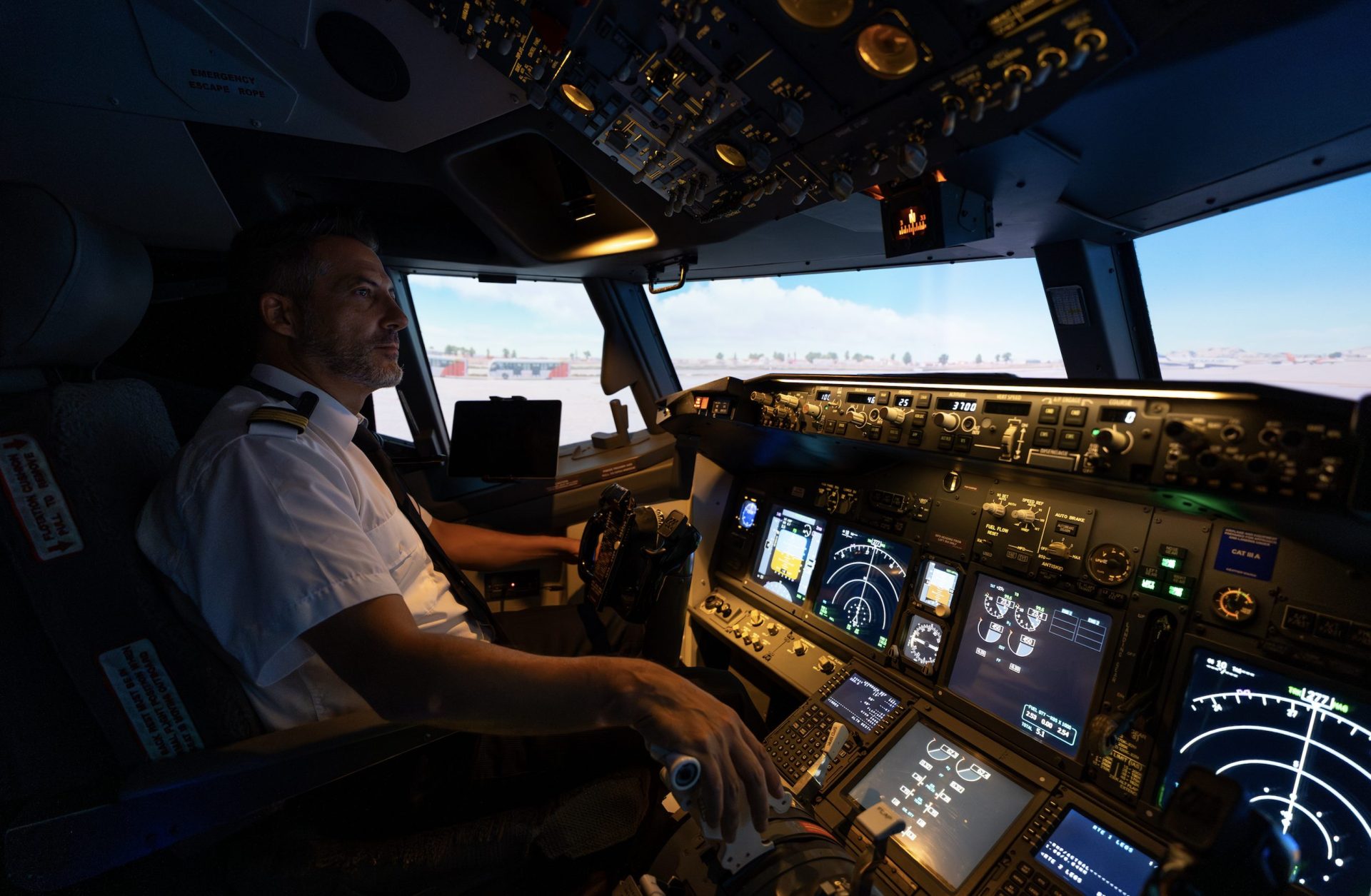 Airline pilot at the controls of a flight simulator, with illuminated cockpit instruments and an airport scene ahead.