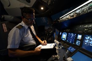 Airline pilot writing on a checklist beside illuminated flight displays in a simulator cockpit.