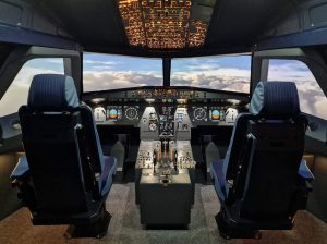 Flight simulator cockpit with two empty pilot seats, illuminated instrument panels and a simulated view above the clouds.